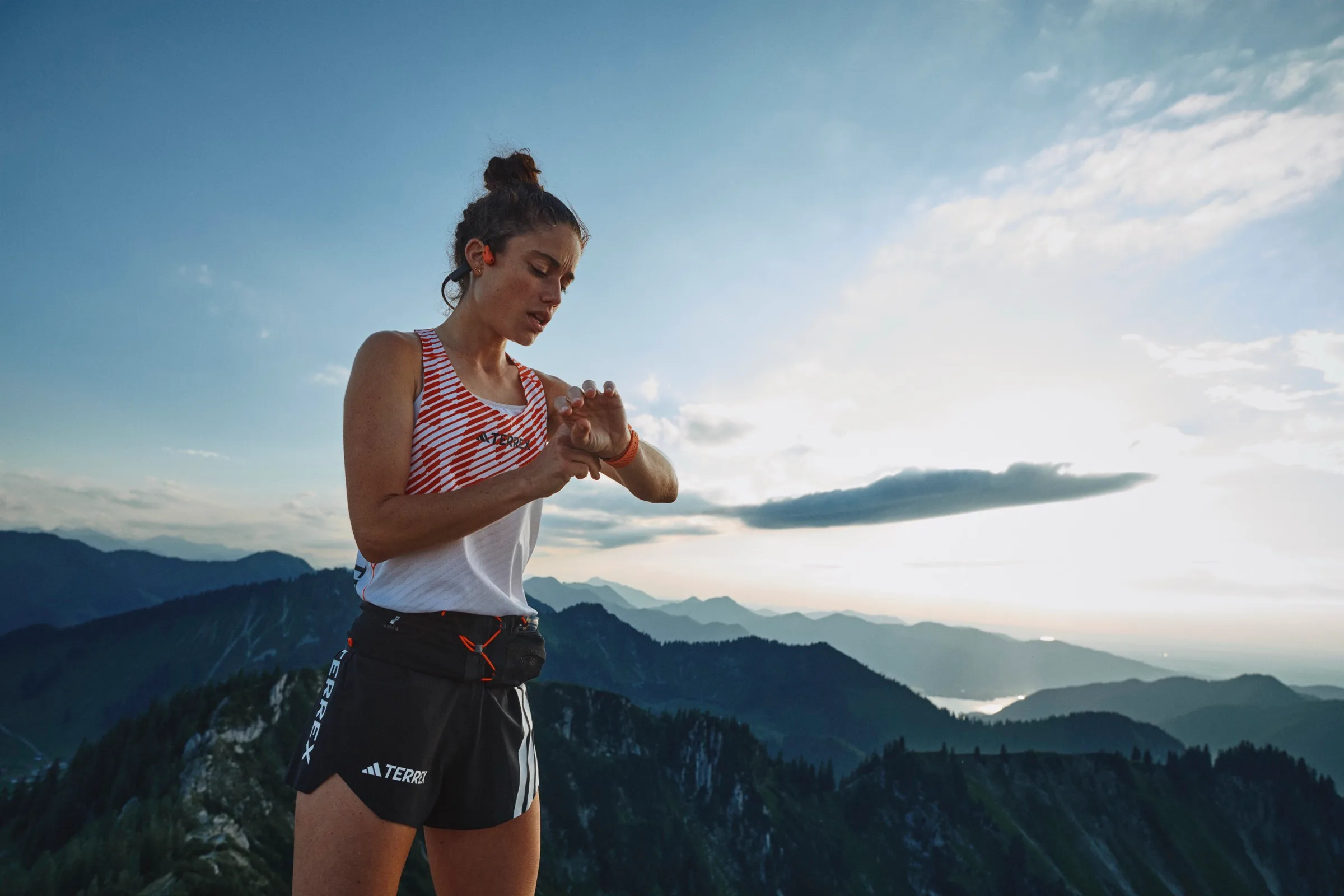 A runner alone on a summit at first light, head down, reading the Suunto on her wrist, with a sea of cloud and receding mountain ridges behind her.